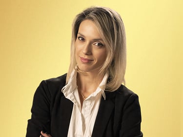 Млад мъж с къса светла коса, облечен в черен пуловер, стои пред жълт фон с кръстосани ръце